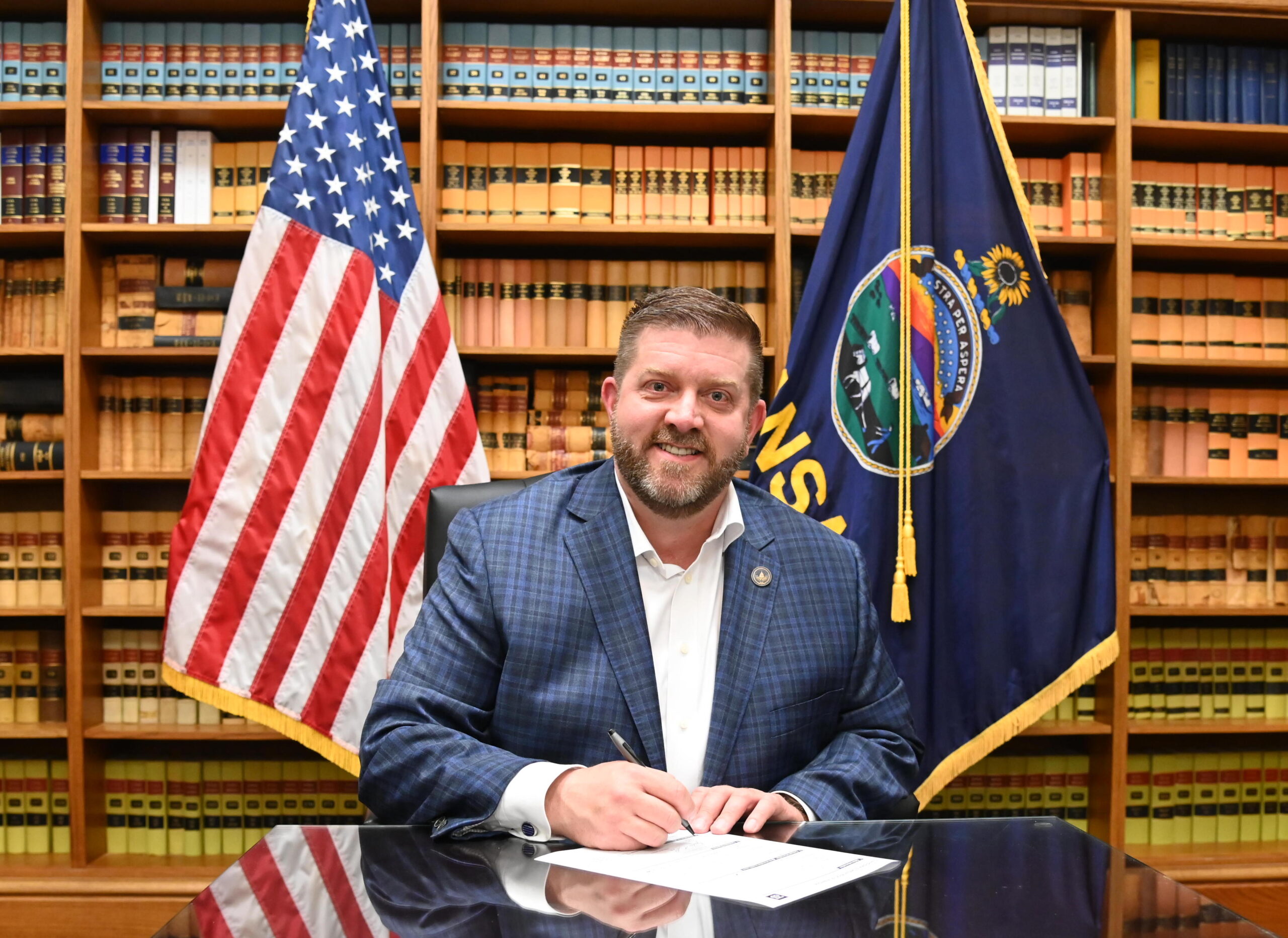 Photo of Justin Shore, signing candidacy paperwork, while seated in front of the US and Kansas flags and bookshelves.